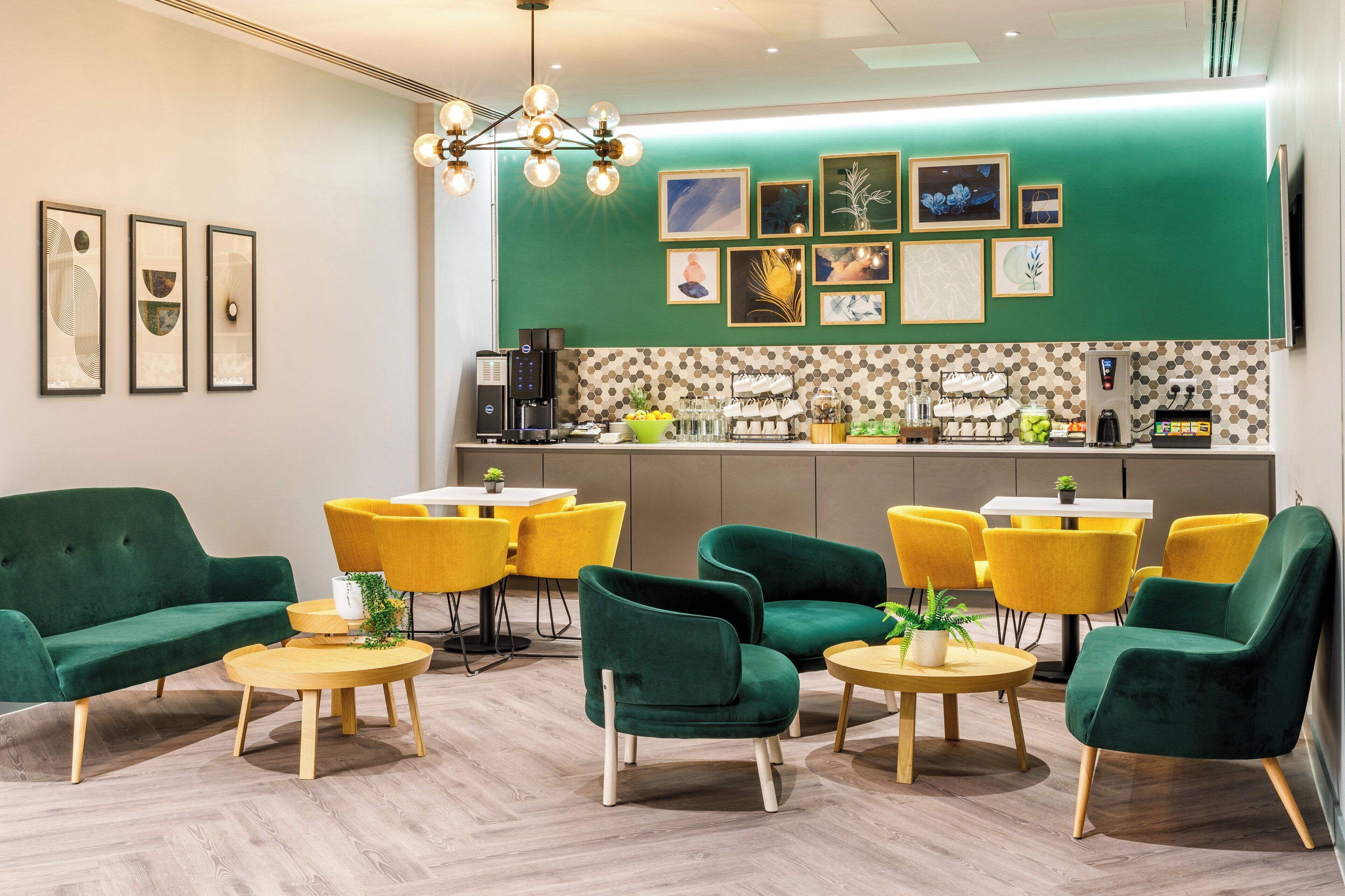 A modern lounge area with green sofas and yellow chairs, a coffee station, and artwork on the wall.