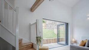 A spacious room with a wooden staircase, a large sliding glass door leading to a garden, and a cozy seating area with a lamp. - Brook Cottage Green - 2 Bedroom Cottage - Dale (Haverfordwest)