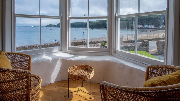 A room with a bay window overlooking a beach, wicker furniture, and a stone wall. - Brook Cottage Green - 2 Bedroom Cottage - Dale (Haverfordwest)