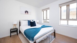 Une chambre à coucher avec un lit, une table de chevet, une lampe, une fenêtre et un tableau encadré au mur.