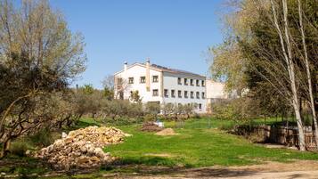 Un edificio grande y blanco con numerosas ventanas, rodeado de árboles y un montículo de piedras.