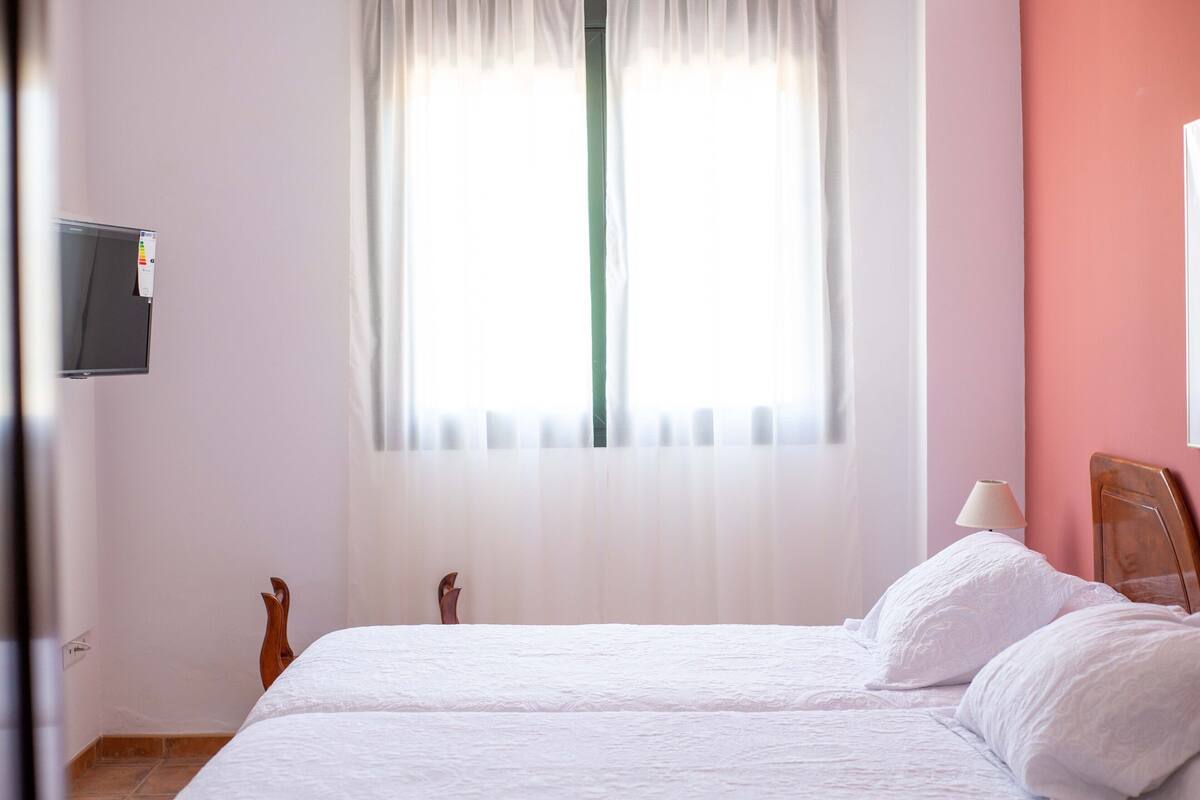 A hotel room with a bed, a TV mounted on the wall, and a window with sheer curtains.