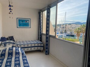 Een kamer met een bed, een ingelijst schilderij, een raam met uitzicht op boten en gebouwen, en een gordijn met anker afbeeldingen.