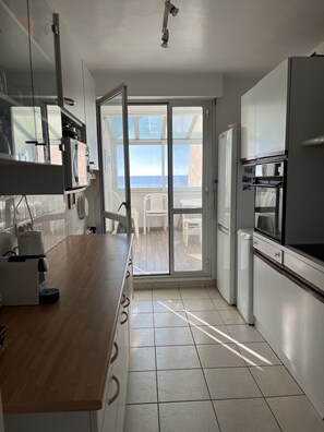 Een moderne keuken met een houten aanrecht, witte kastjes en uitzicht op zee door een glazen deur.