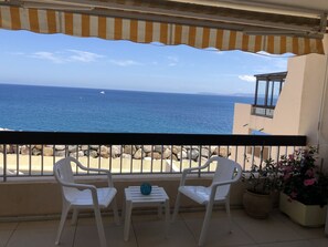 Een balkon met twee witte plastic stoelen, een kleine tafel met een blauwe vaas en een potplant met roze bloemen. De horizon toont de zee en een boot in de verte.