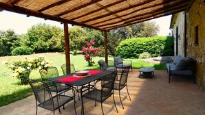 An outdoor patio with a table and chairs, a covered area with a thatched roof, and a garden with flowers and greenery. - I run away to Umbria, Monti (Umbria)