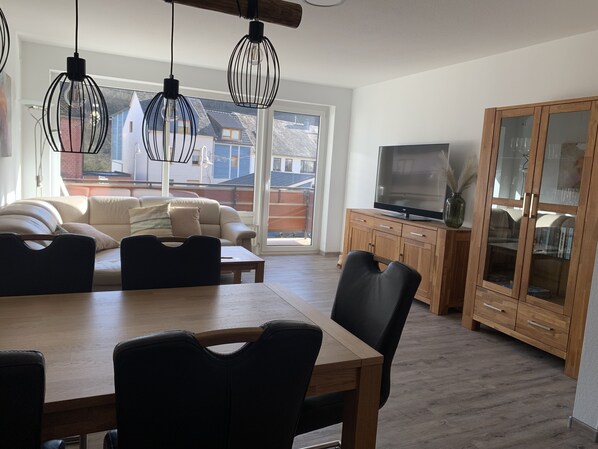 A spacious living room with a wooden dining table, black chairs, a sofa, and a TV on a wooden cabinet.