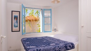 A bedroom with a bed, a window with blue shutters, a framed picture on the wall, and a hanging light fixture.