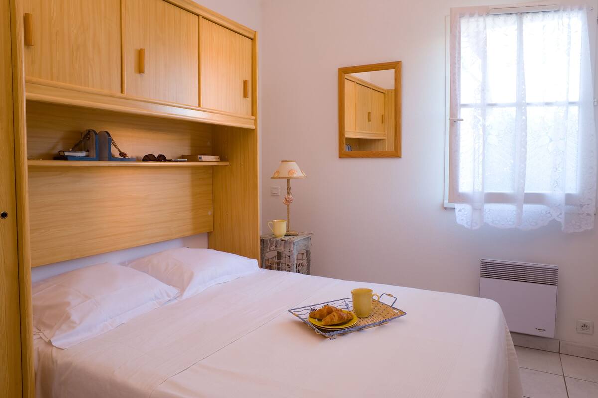 A neatly made bed with a tray of croissants and a cup, wooden headboard, a mirror, and a window with lace curtains.
