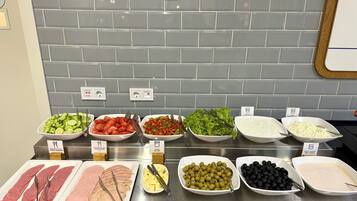 A buffet with various food items including sliced cucumbers, tomatoes, lettuce, butter, fruit yogurt, olives, black olives, cheese, ham, and turkey.