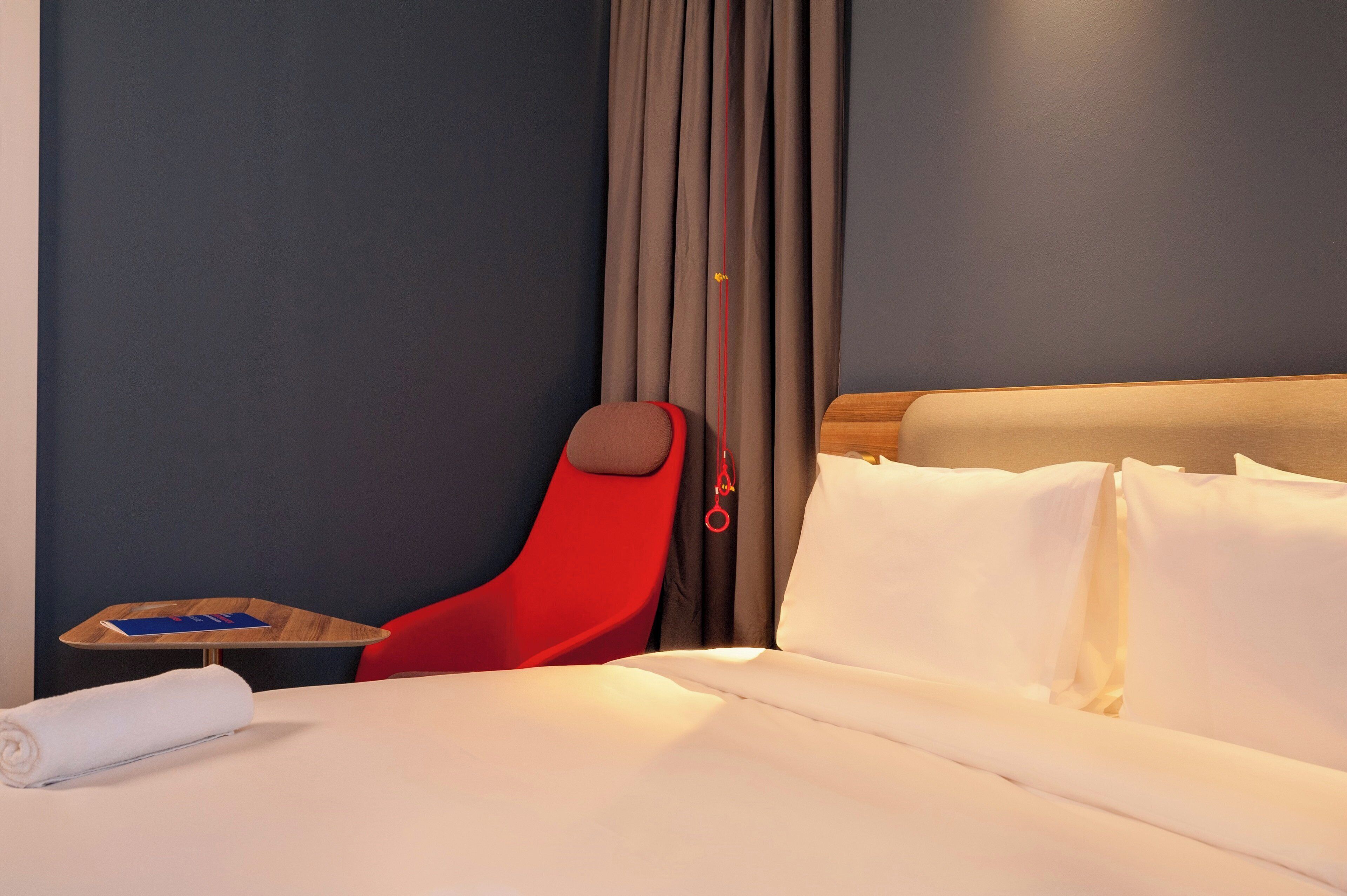 Une chambre d’hôtel avec un lit, une chaise rouge, une table de chevet et un rideau.