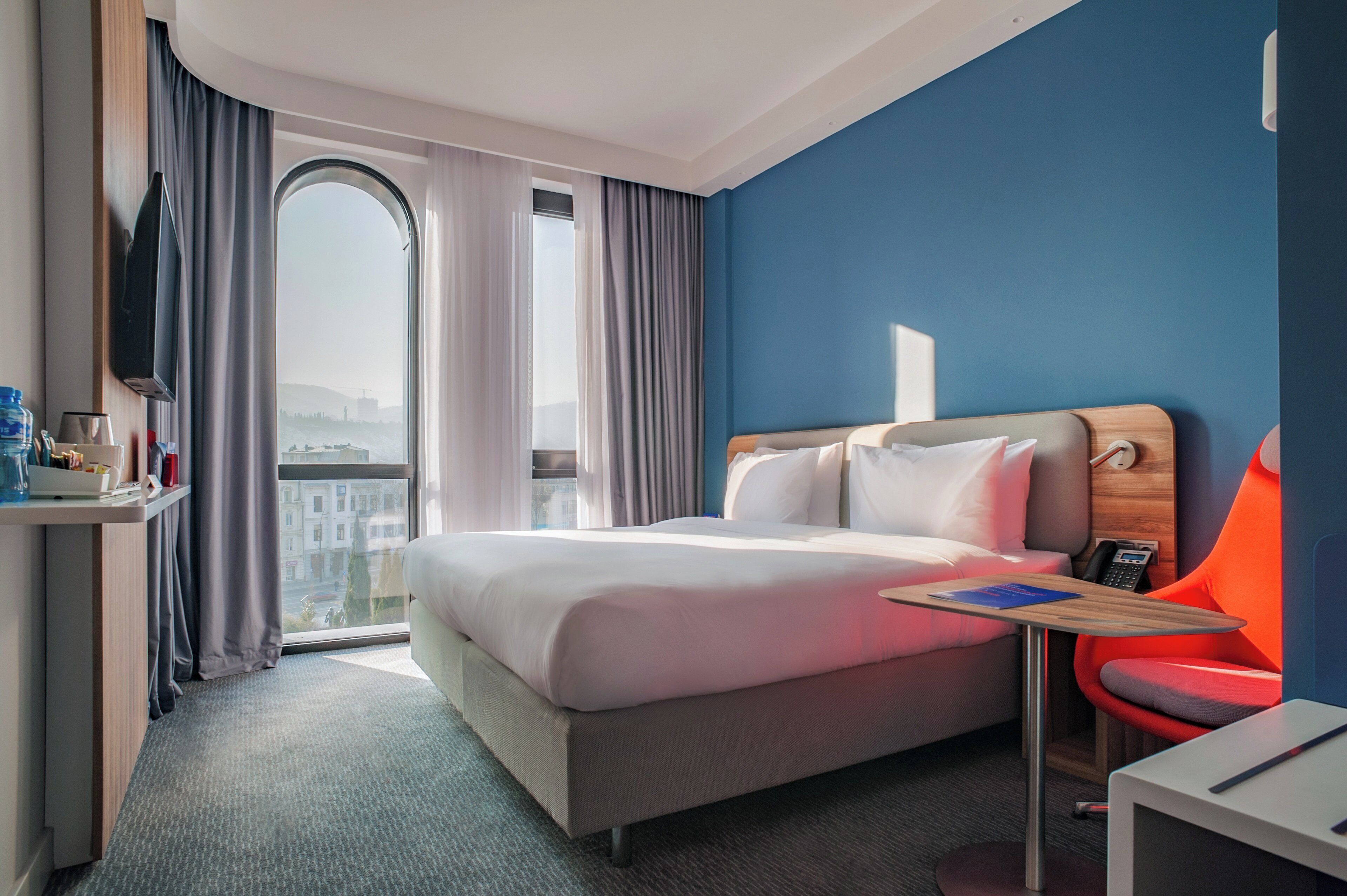 A hotel room with a bed, a desk, a chair, a TV, and a window with curtains.