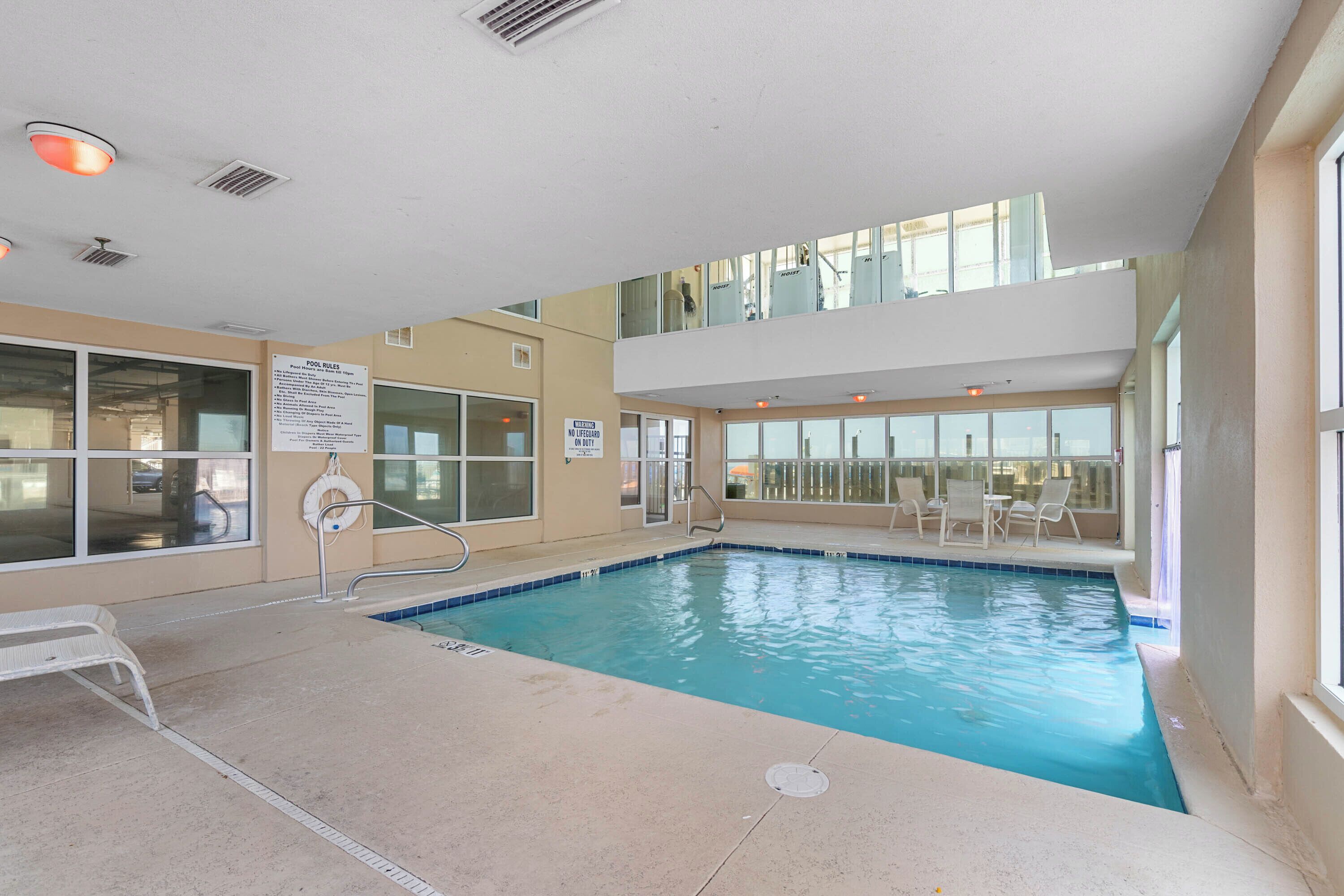 An indoor swimming pool with clear pool rules posted, a lifeguard sign, and a view of the city through large windows.