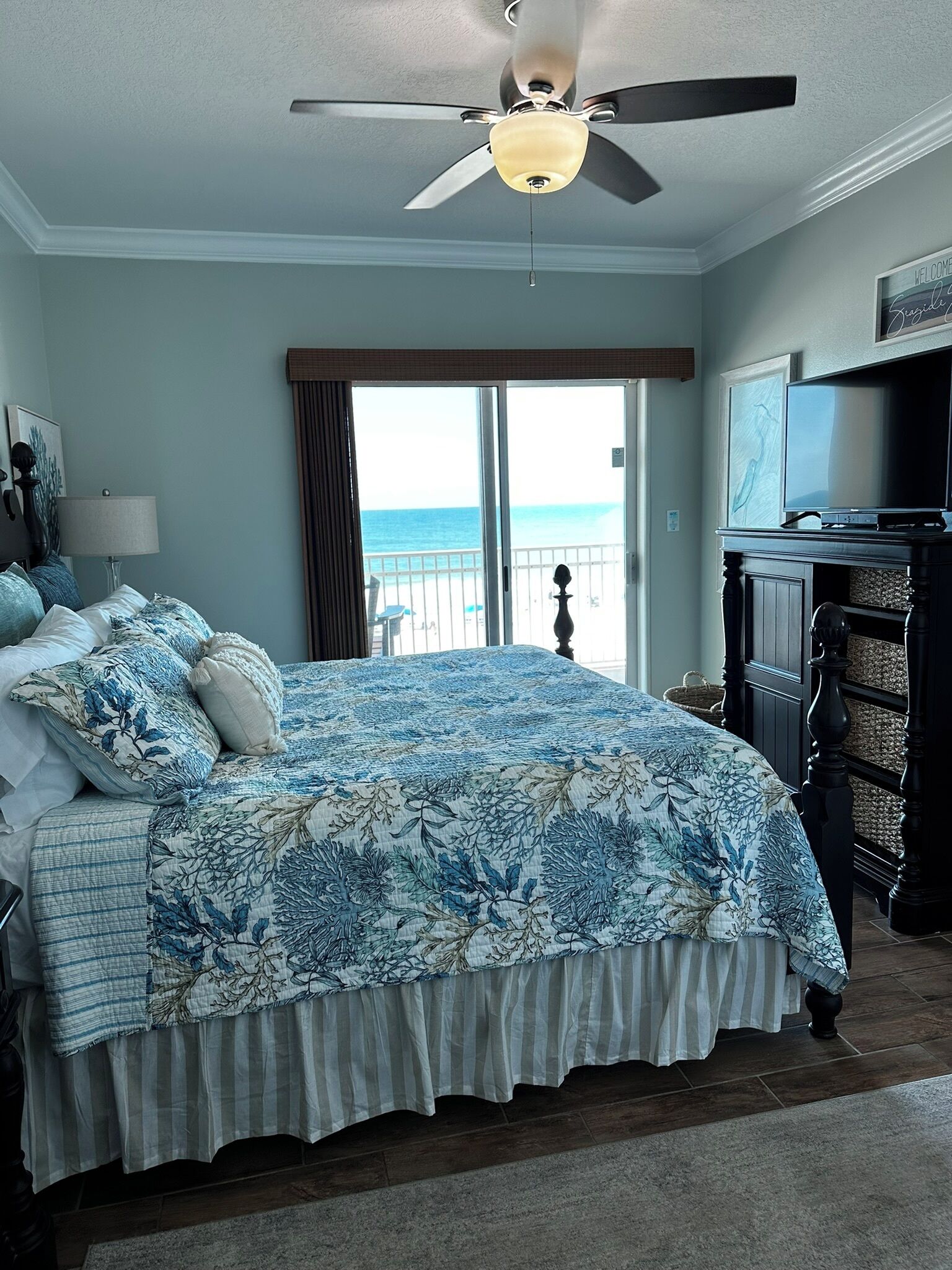 A bedroom with a large bed, a ceiling fan, a TV, and a view of the ocean.