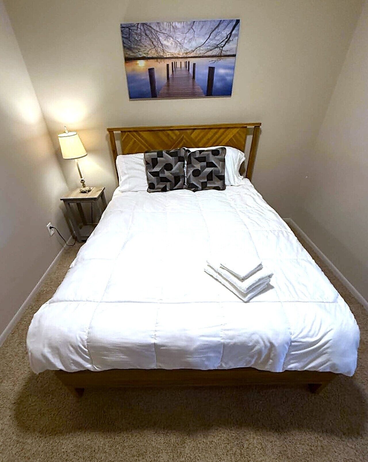 Una cama bien hecha con ropa blanca, cabecera de madera, lámpara de noche y un cuadro de un puente sobre el agua en la pared.