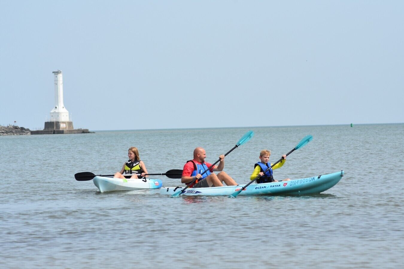 Trois personnes pratiquent le kayak sur une eau calme, avec un phare en arrière-plan.