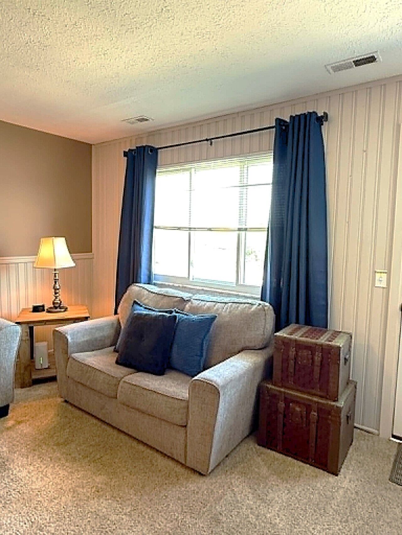 A living room with a beige sofa, two blue cushions, a lamp, a window with blue curtains, and a wooden chest of drawers.