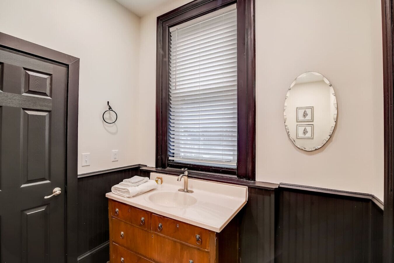 Un baño con puerta oscura, una ventana con persianas, un lavabo con espejo y un perchero para toallas.