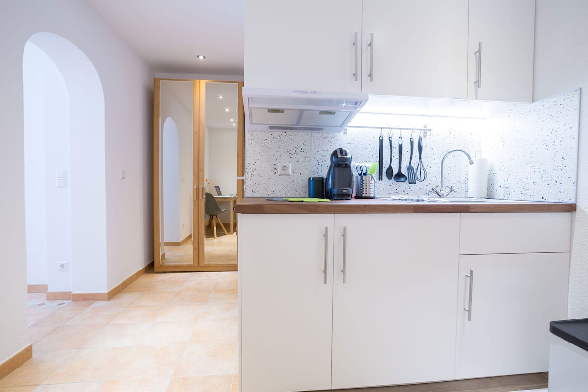 A modern kitchen with white cabinets, a countertop, and a coffee maker. There is a door leading to another room.