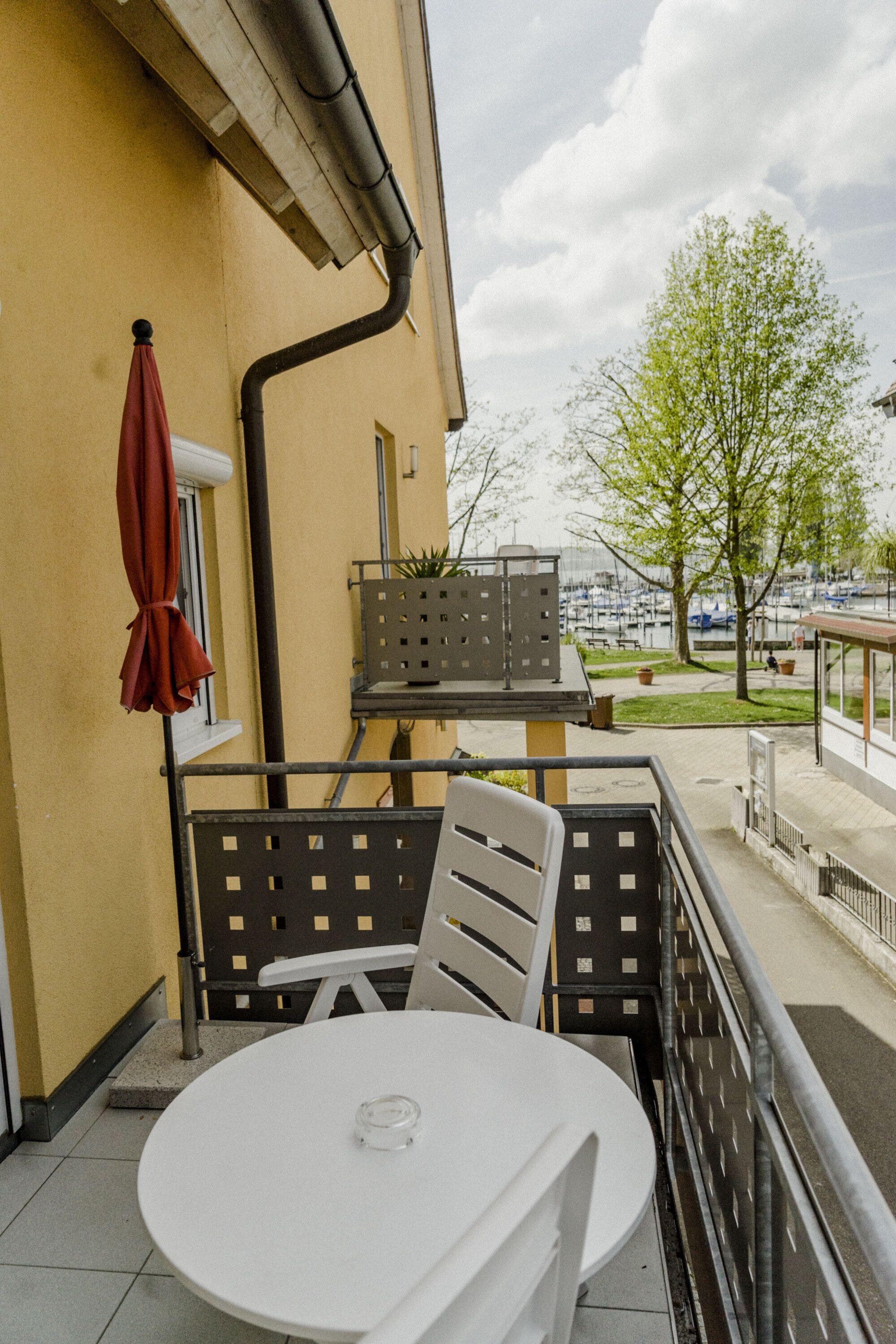 A balcony with a white table and chairs, a red umbrella, and a view of a marina with boats.