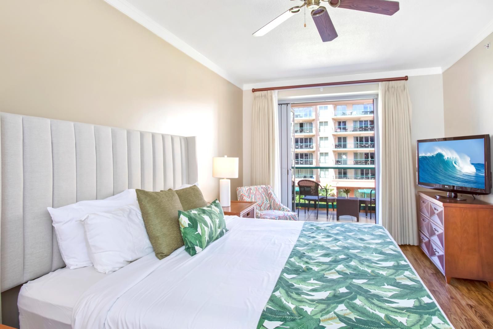 A hotel room with a bed, a television, a balcony with a view of the city, and a ceiling fan.