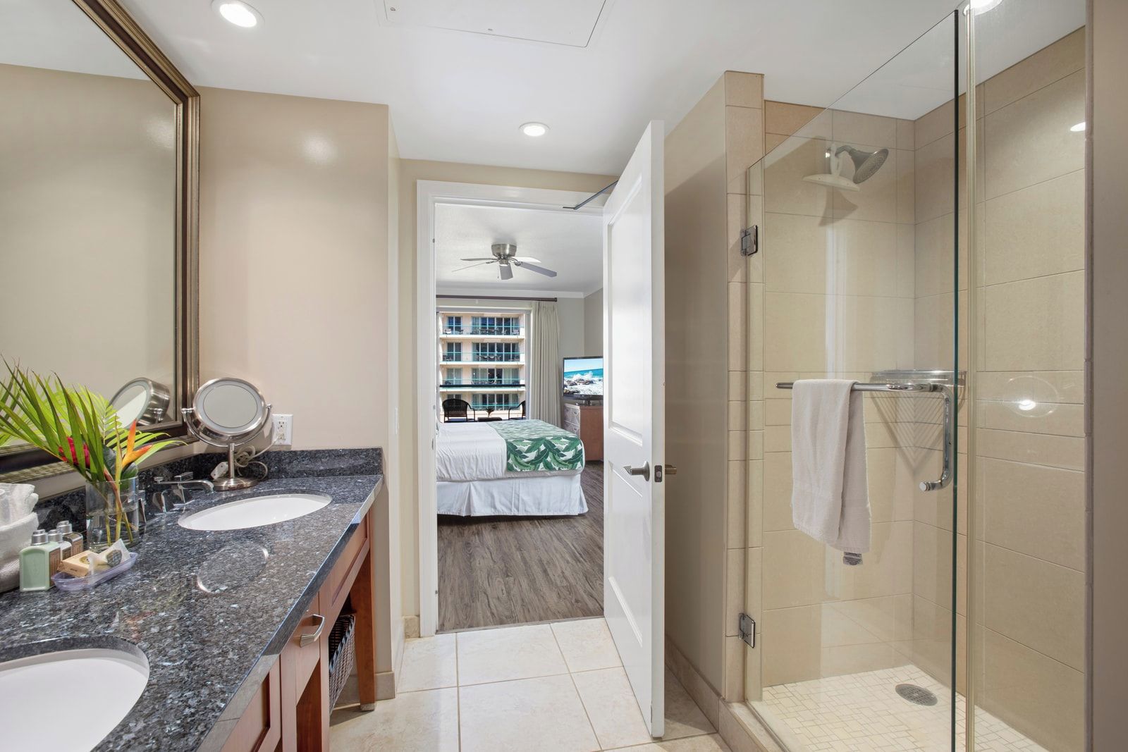 Une salle de bain d’hôtel avec un double lavabo, une douche et un lit visible à travers une porte ouverte.