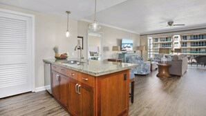 A spacious living area with a large window, a kitchen with granite countertops, and a ceiling fan. - Honua Kai | Ocean View 2 BR Sleeps 8 | Car Incl. w/ 6+ Nights | HKK-309 by KBM (Lahaina)