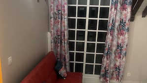 Ein rotes Sofa mit gemusterten Kissen in einem Raum mit Fliesenboden und einer Glastür mit floralen Vorhängen.