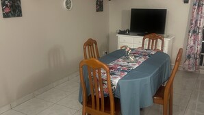 Ein Essbereich mit einem Tisch für vier Personen, Holzstühle, einem Fernseher auf einem weißen Schrank und einem gefliesten Boden.