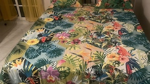 Ein Bett mit Bettdecke mit floralem und tropischen Muster, einem Holzkopfschild und einem gelben Vorhang.