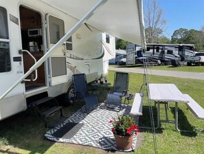 A recreational vehicle with a covered patio area, two chairs, a table, and a potted plant. - Vacation RV Rental Near Disney with Pool (Kissimmee)