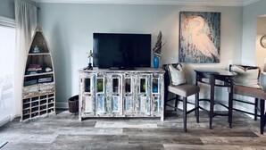 A living room with a TV on a wooden cabinet, a bookshelf, a painting, and a dining area with chairs and a table. - 1BR/1.5BA retreat with direct beach access (Orange Beach)