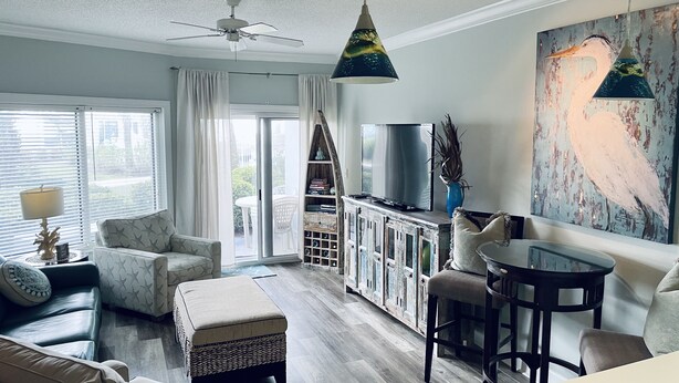 A modern living room with a sofa, ottoman, and a TV on a wooden cabinet. There is a ceiling fan, a dining area with a table and chairs, and a large window with curtains. - 1BR/1.5BA retreat with direct beach access (Orange Beach)