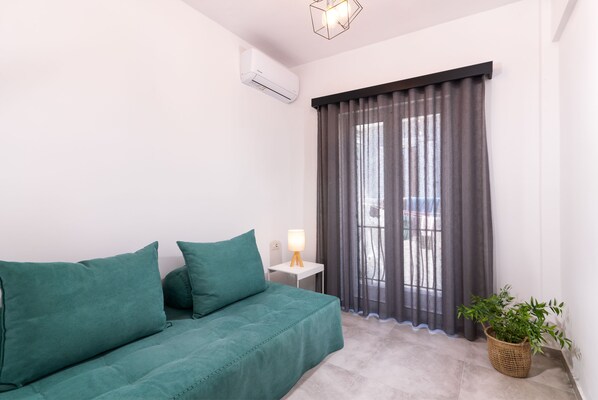 Una habitación con un sofá verde, una mesita de noche con lámpara, una planta en maceta y una ventana con cortinas grises.
