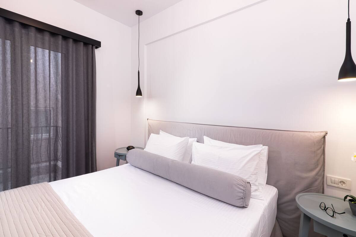 A modern hotel room with a large bed, a bedside table with a lamp, and a view of the outdoors through a window with curtains.