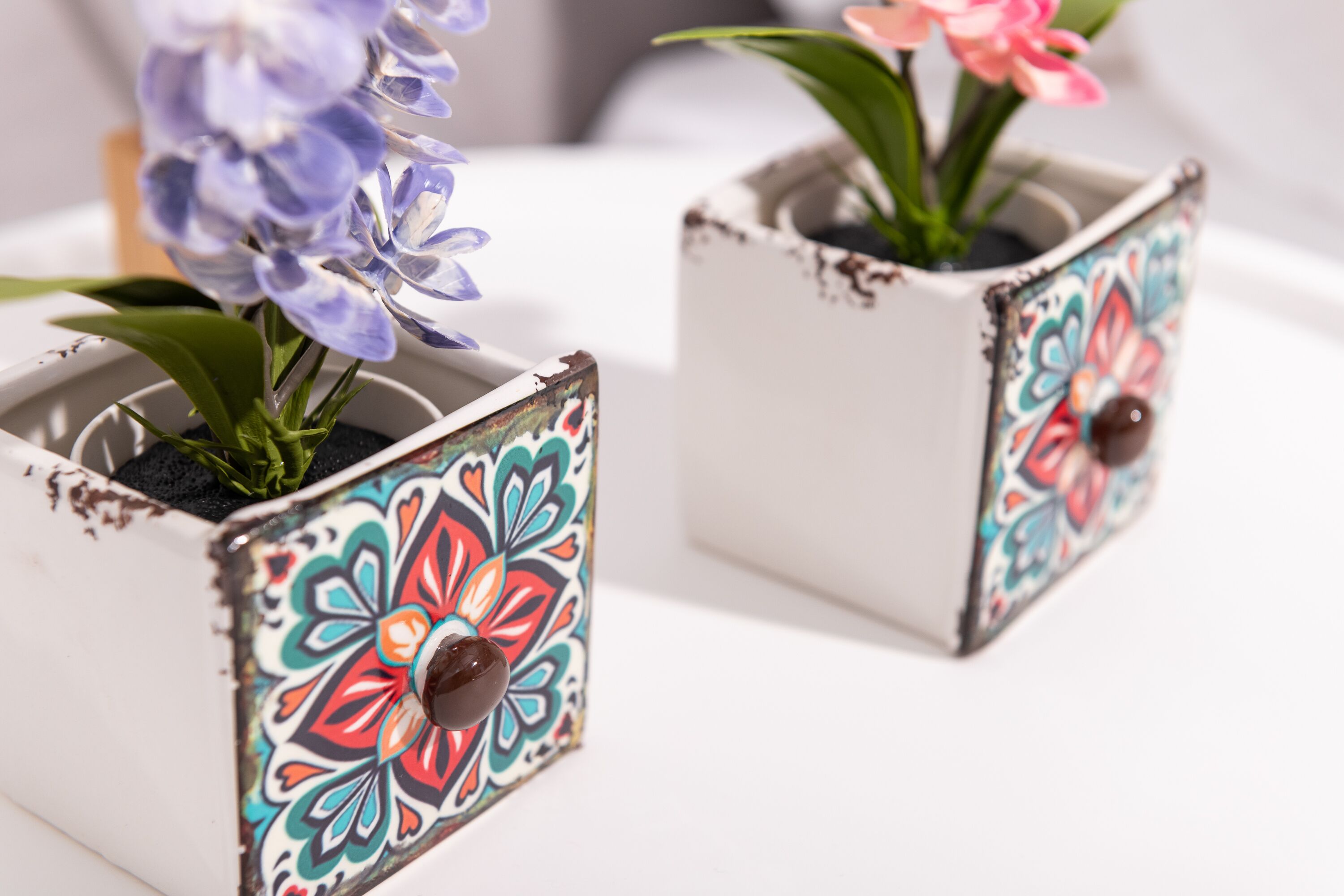 Deux jardinières carrées ornées de motifs floraux colorés, chacune surmontée d’un bouton marron.