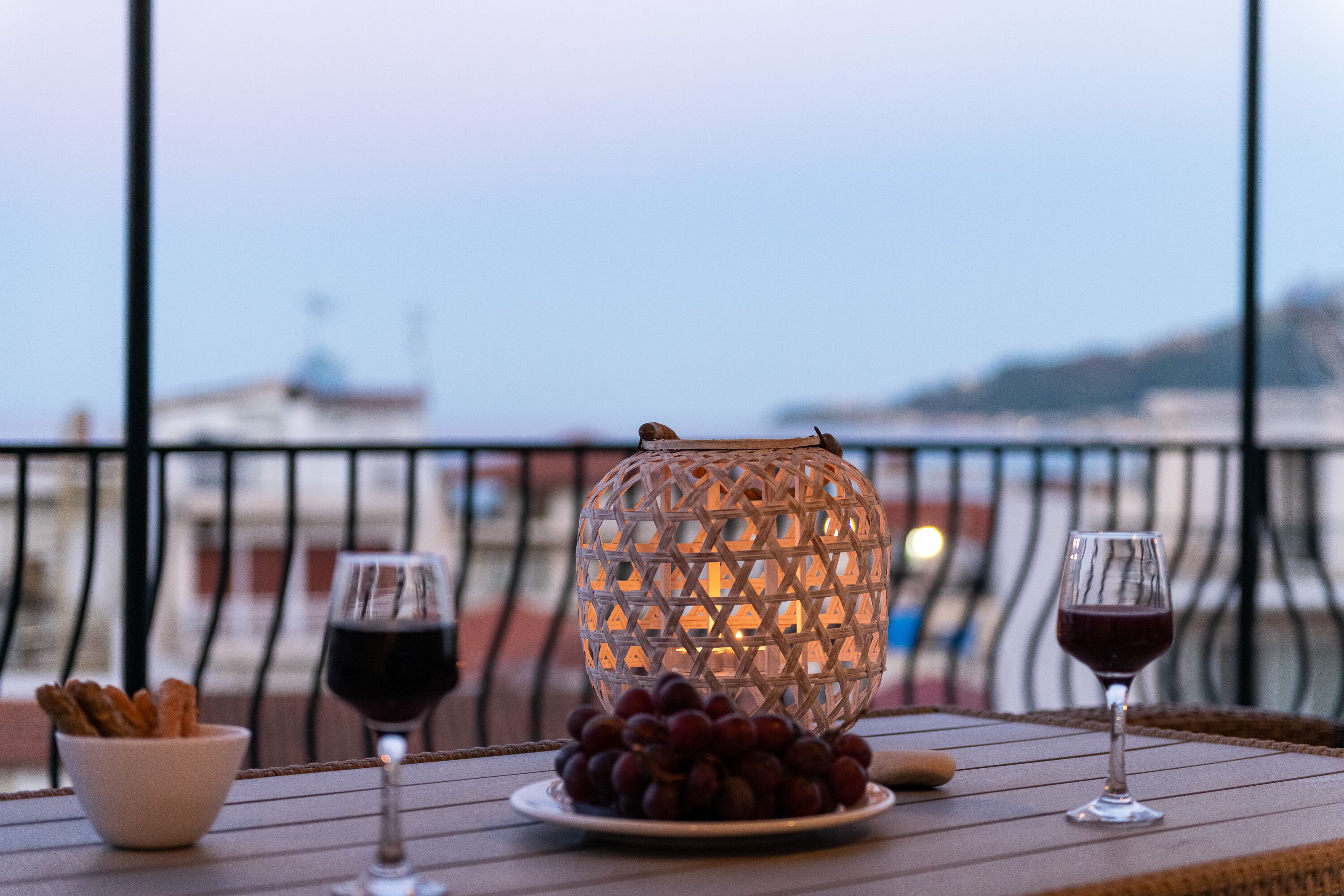 Une table avec un verre de vin rouge, un bol de raisins et une lanterne décorative en verre, le tout sur un balcon donnant sur une vue panoramique de la ville.