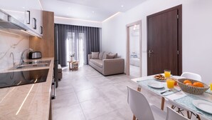 A modern kitchen with a built-in oven, a dining area with a table set for breakfast, and a living area with a sofa and a view of the city. - Lithies Zante Living (Zakynthos)