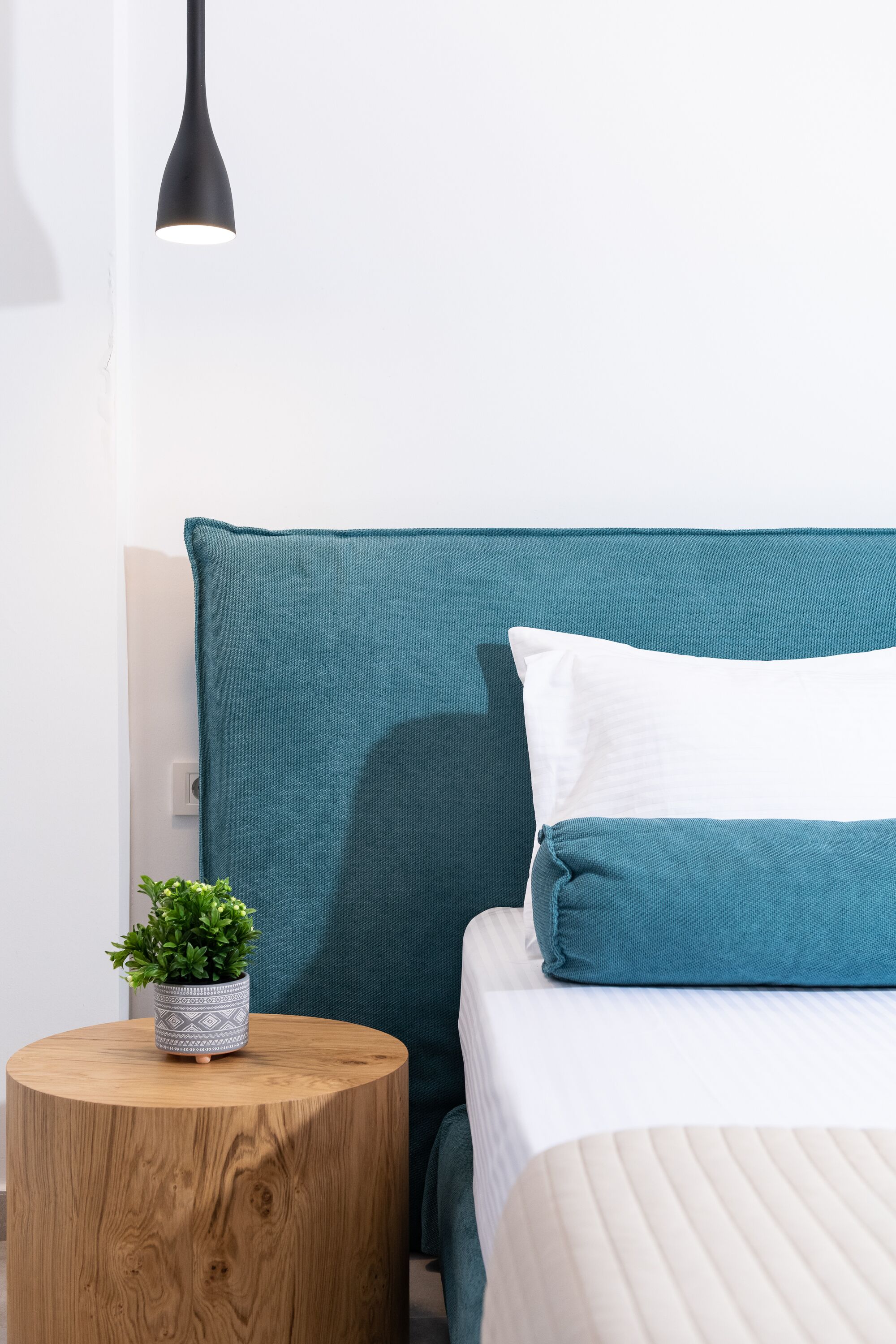Una cama bien hecha con ropa de cama color turquesa y una almohada blanca, una pequeña mesita de madera con una planta y una lámpara colgante negra.