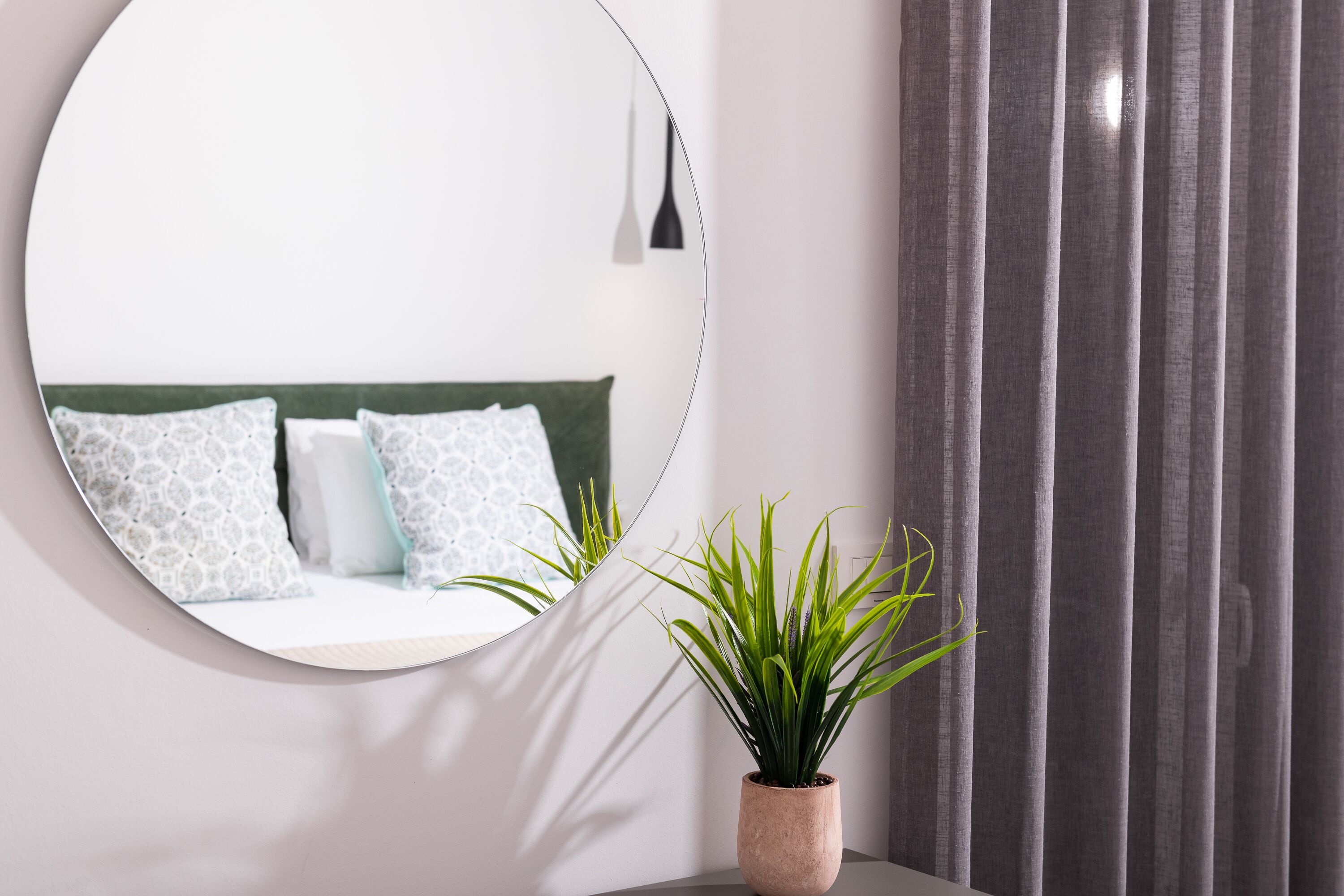 A round mirror reflecting a bed with patterned pillows and a green headboard. A potted plant is in front of the mirror.