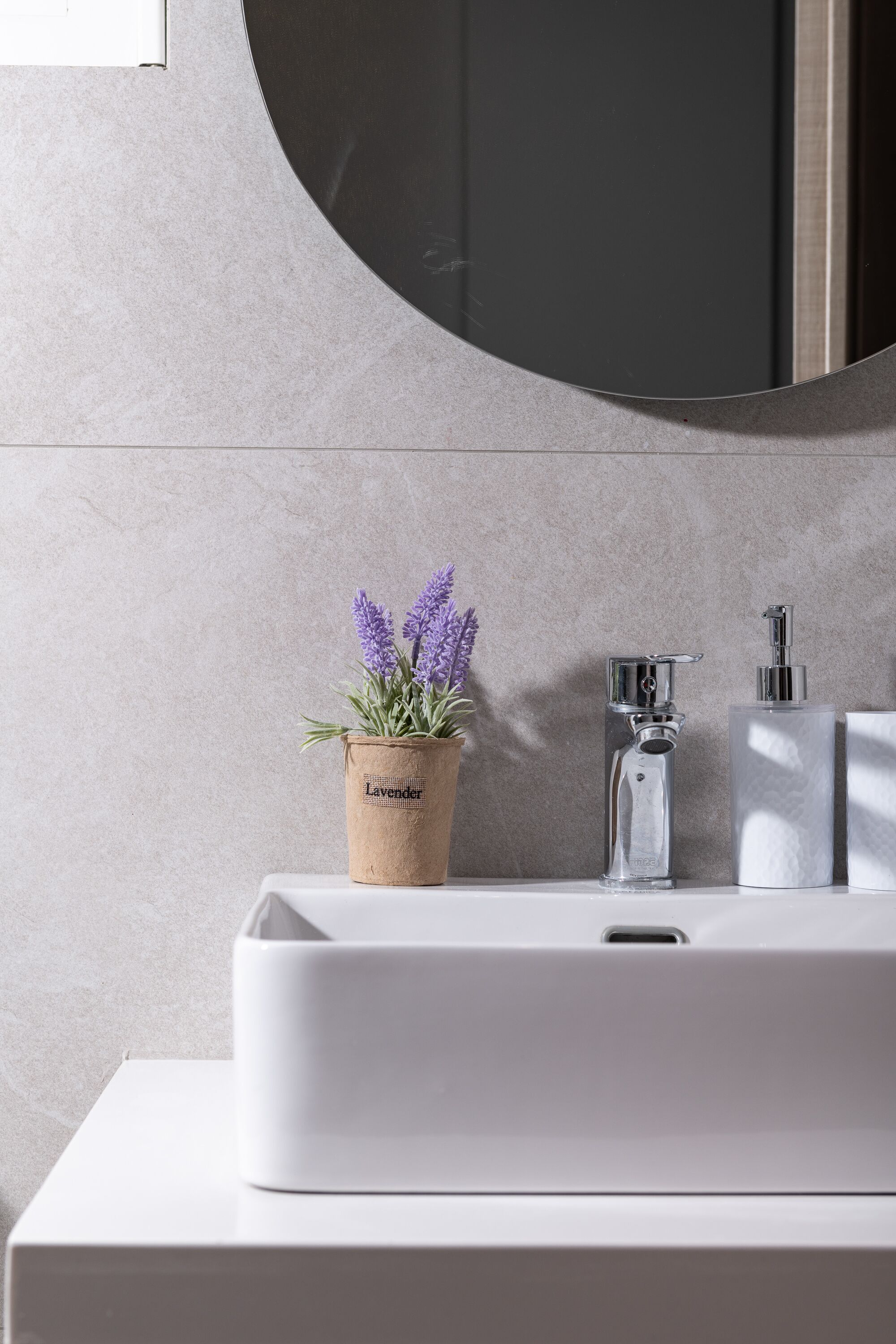 Un baño con un lavabo blanco, un espejo redondo y una planta de lavanda en una maceta marrón.