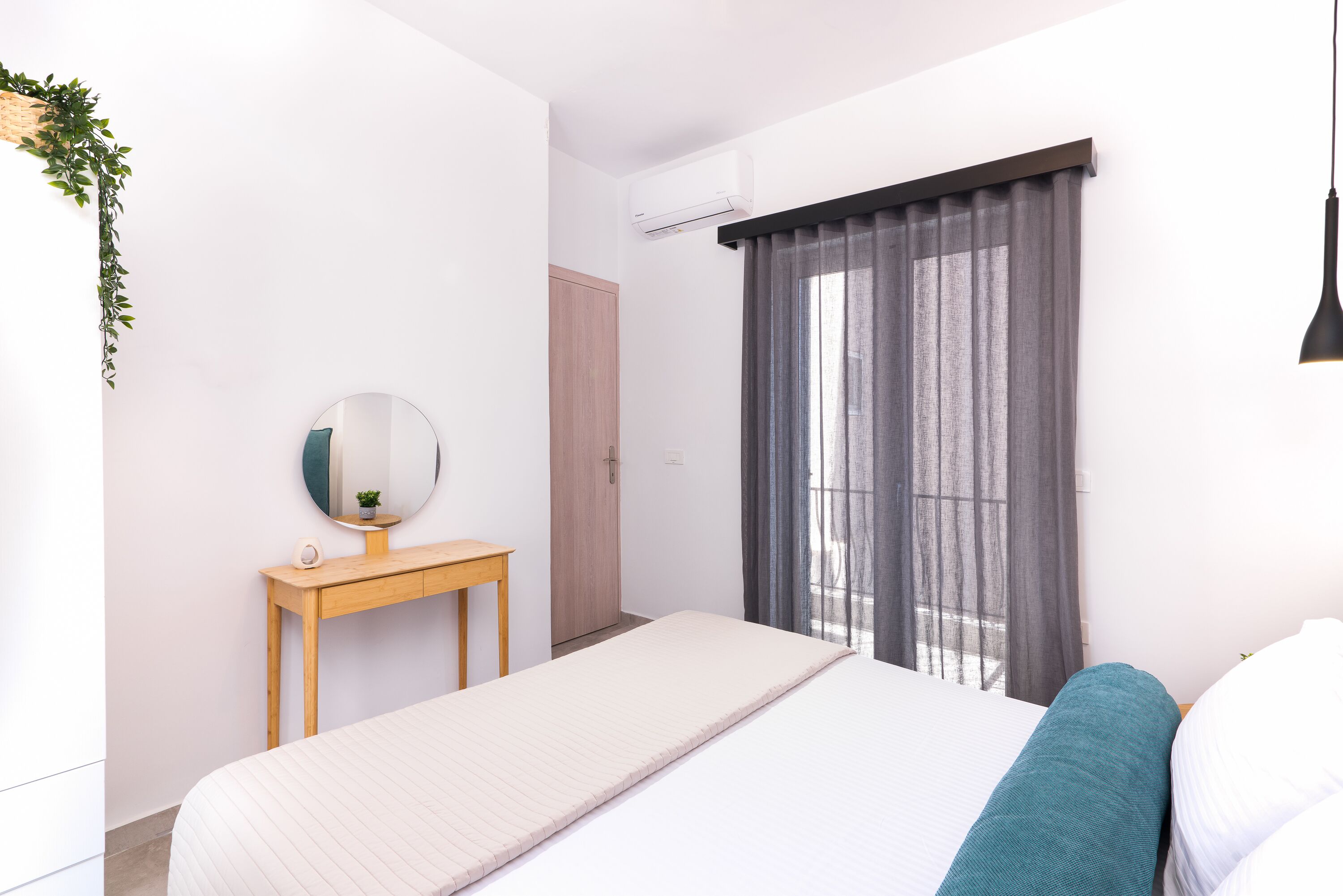 Une chambre à coucher minimaliste avec un lit, une table de chevet dotée d’un miroir, une petite plante et une fenêtre avec des rideaux.