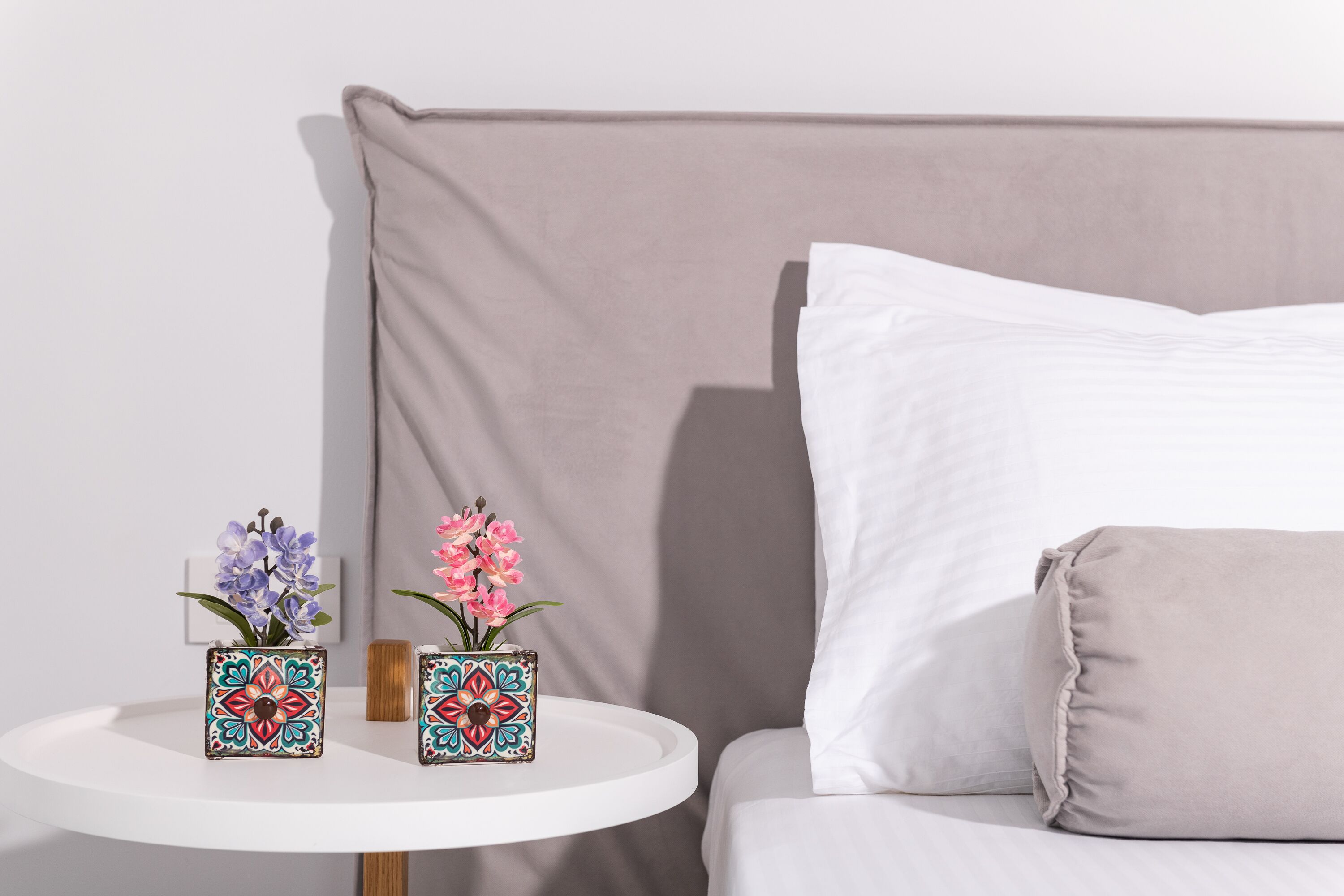 Una cama con cabecera color crema y almohadas, una mesita blanca pequeña con dos jarrones decorativos y una mesita de noche de madera.