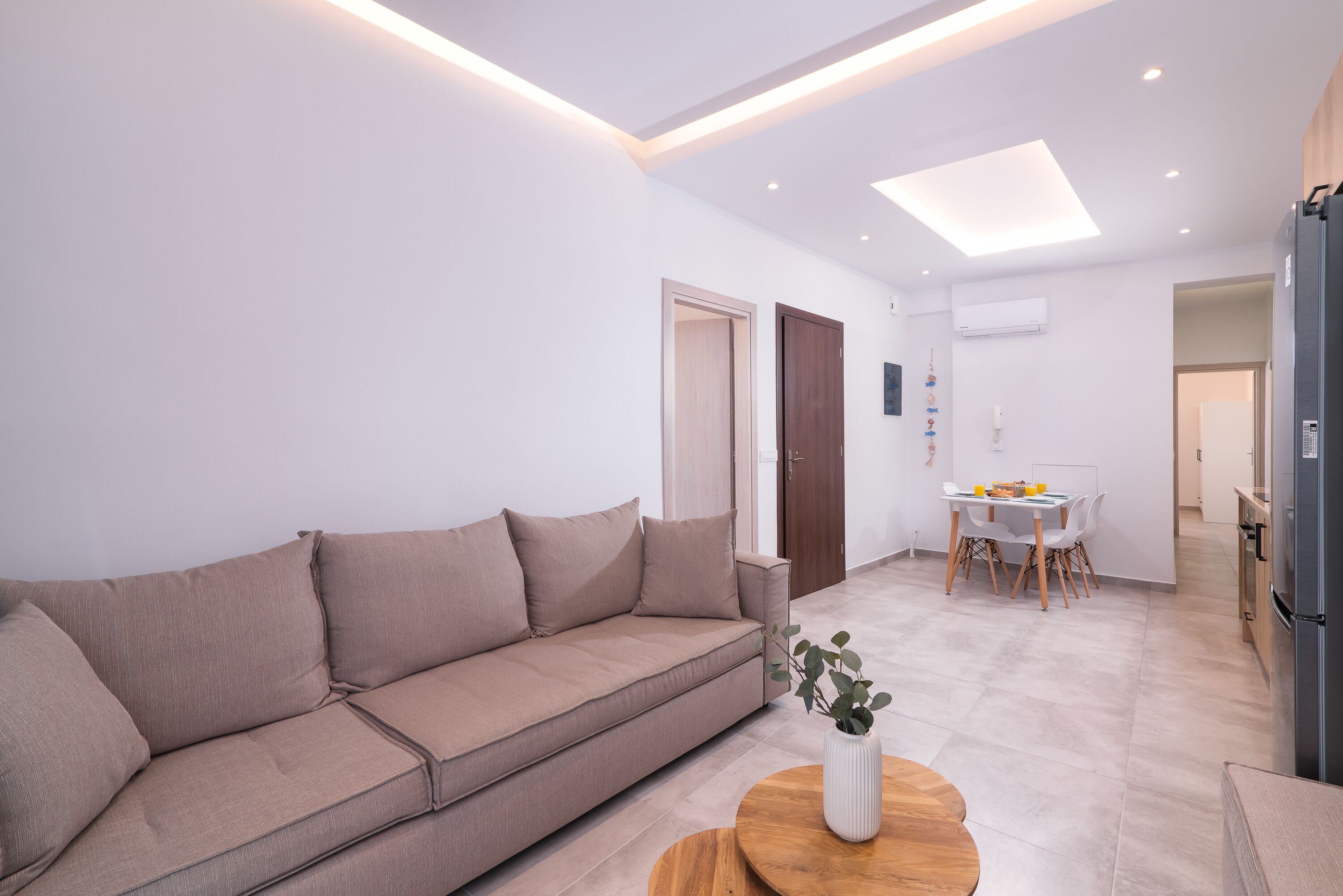 Una sala de estar moderna con un sofá color beige, una mesa de centro de madera y un comedor con una mesa y sillas blancas.