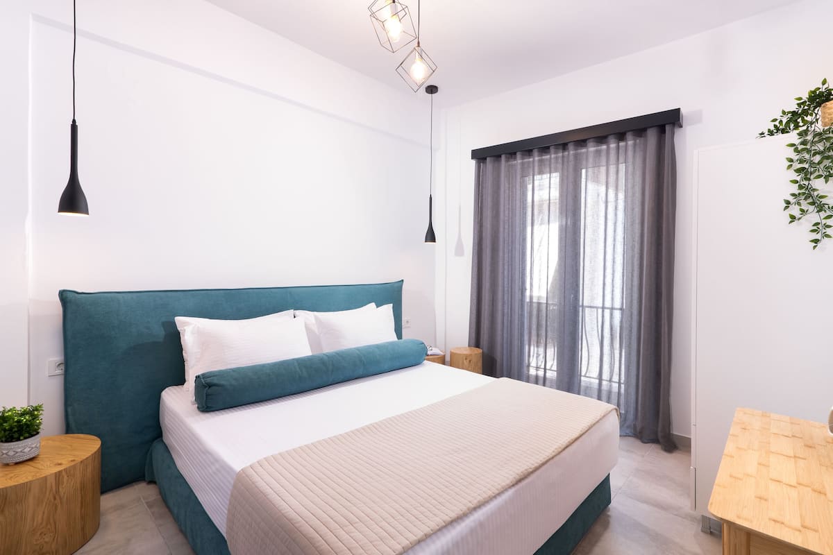 A modern bedroom with a teal headboard, a bed with white and teal bedding, a bedside table with a plant, and a window with grey curtains.