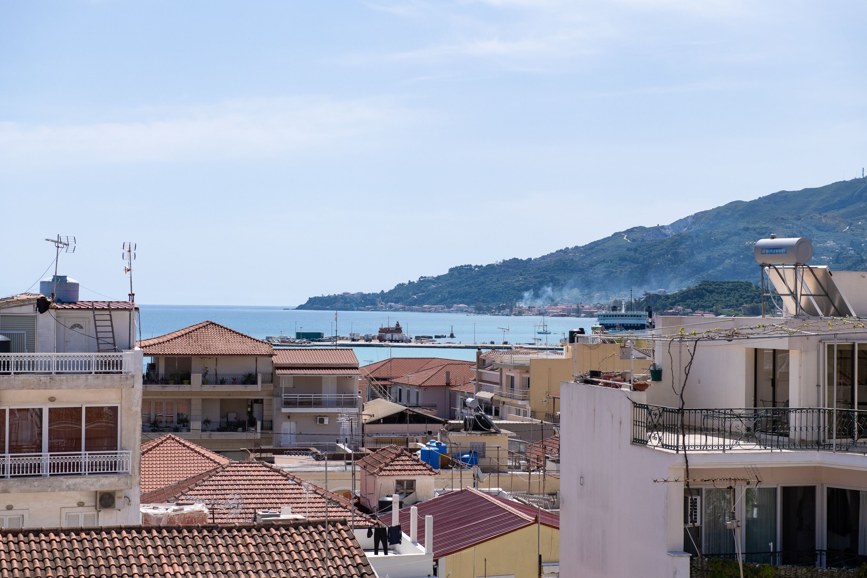 A coastal residential area with terracotta-tiled roofs, balconies, and a view of the sea and distant hills.