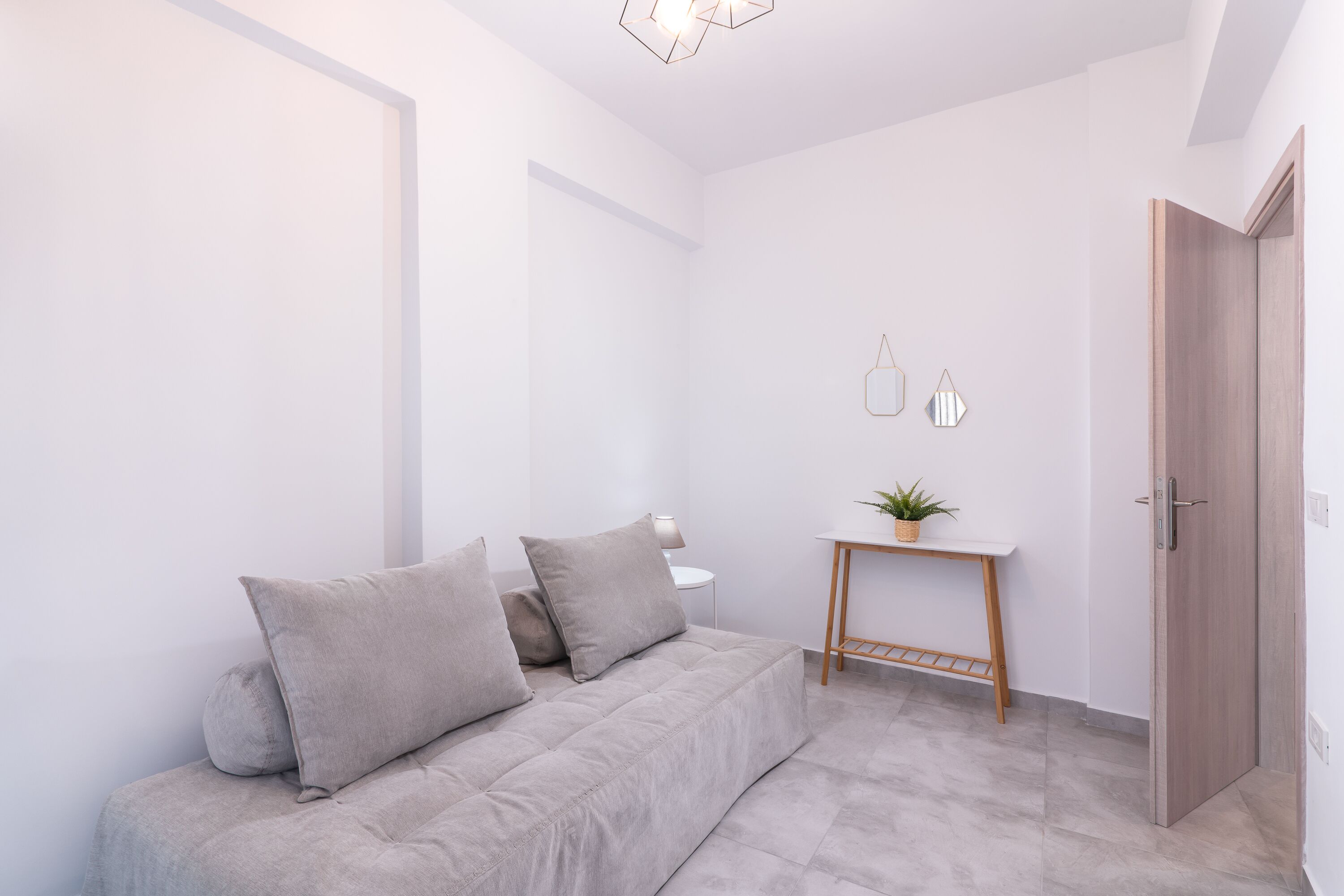 A modern, minimalist living room with a grey sofa, a small wooden table with a plant, and a door.