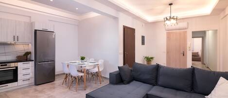 Une cuisine moderne dotée d’un four et d’un réfrigérateur encastrés, un coin repas avec une table et des chaises blanches, et un canapé d’angle gris foncé.