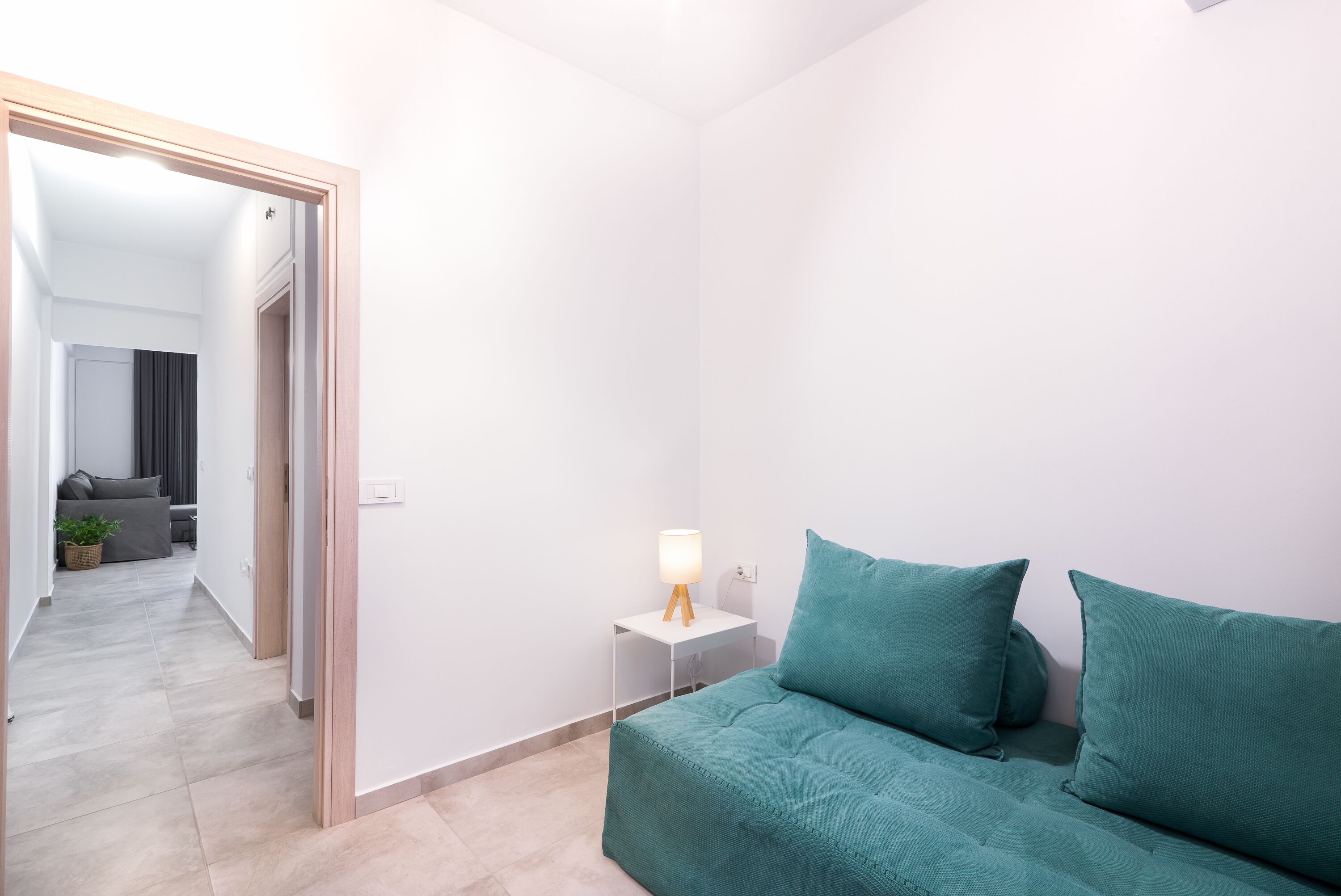 A modern, minimalistic room with a teal sofa, a small bedside table with a lamp, and a view into another room with a grey sofa and a plant.
