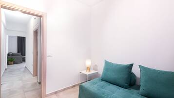 A modern, minimalistic room with a teal sofa, a small bedside table with a lamp, and a view into another room with a grey sofa and a plant.